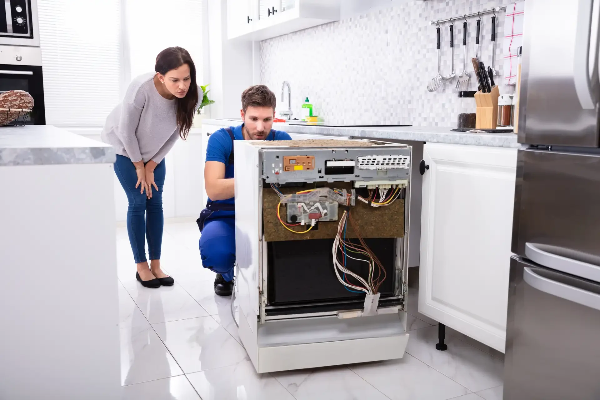 Professional technician repairing dishwasher with front panel removed, exposing internal wiring and control board, while homeowner observes in modern kitchen