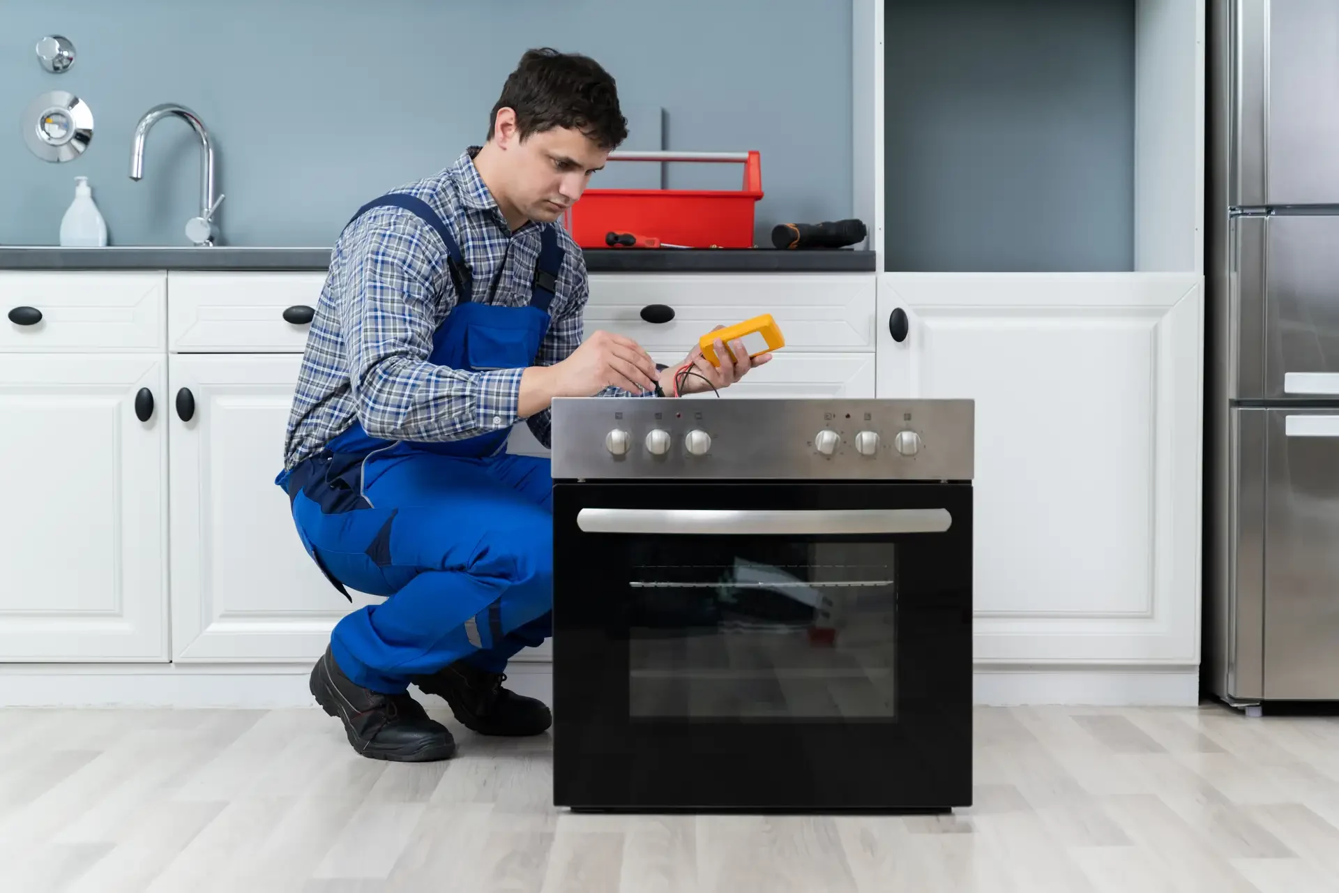 Appliance repair technician in blue overalls using digital multimeter to test electrical components on built-in stainless steel oven with toolbox nearby