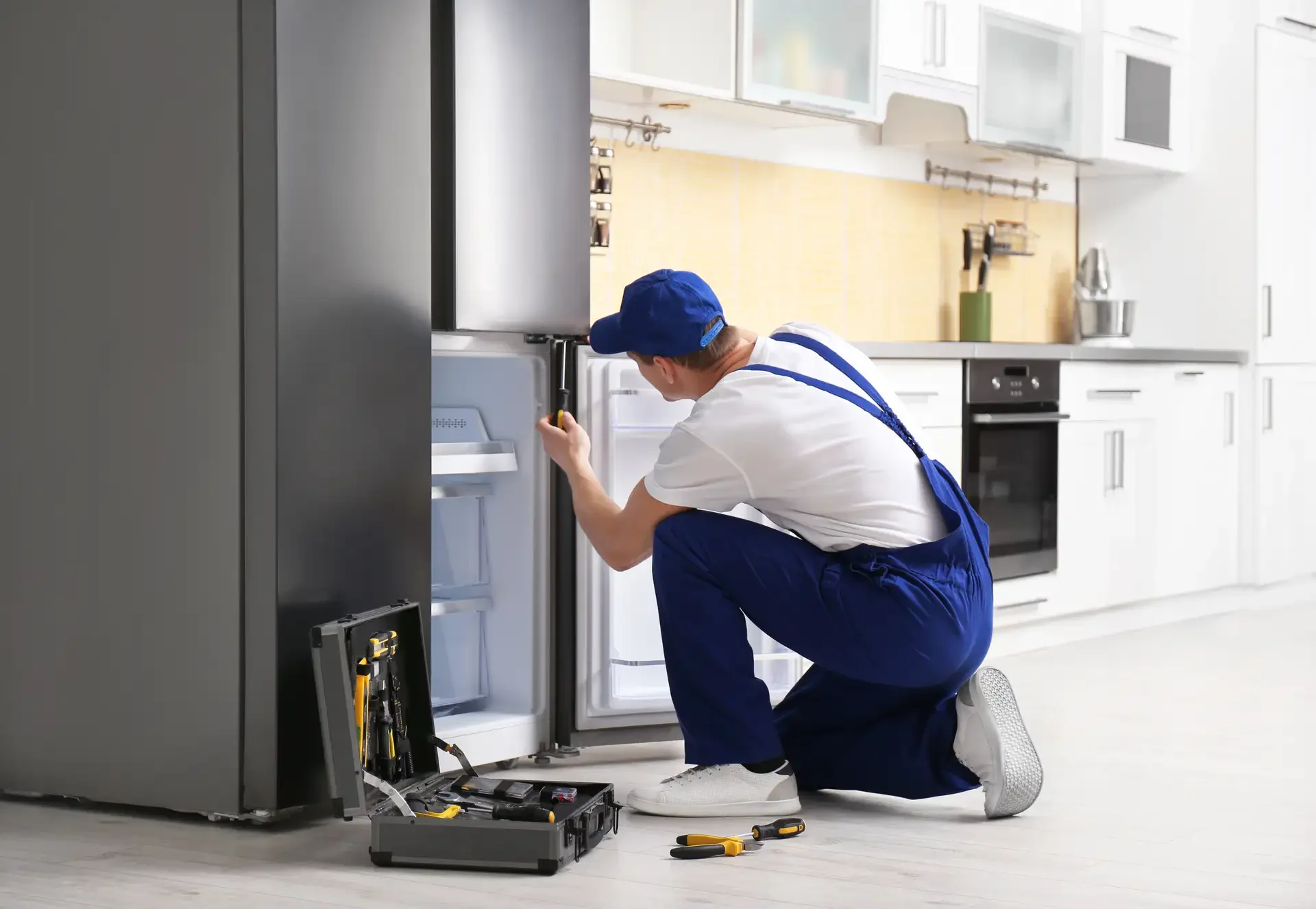 Professional technician in blue uniform repairing an open refrigerator door with screwdriver, toolkit with tools visible on kitchen floor