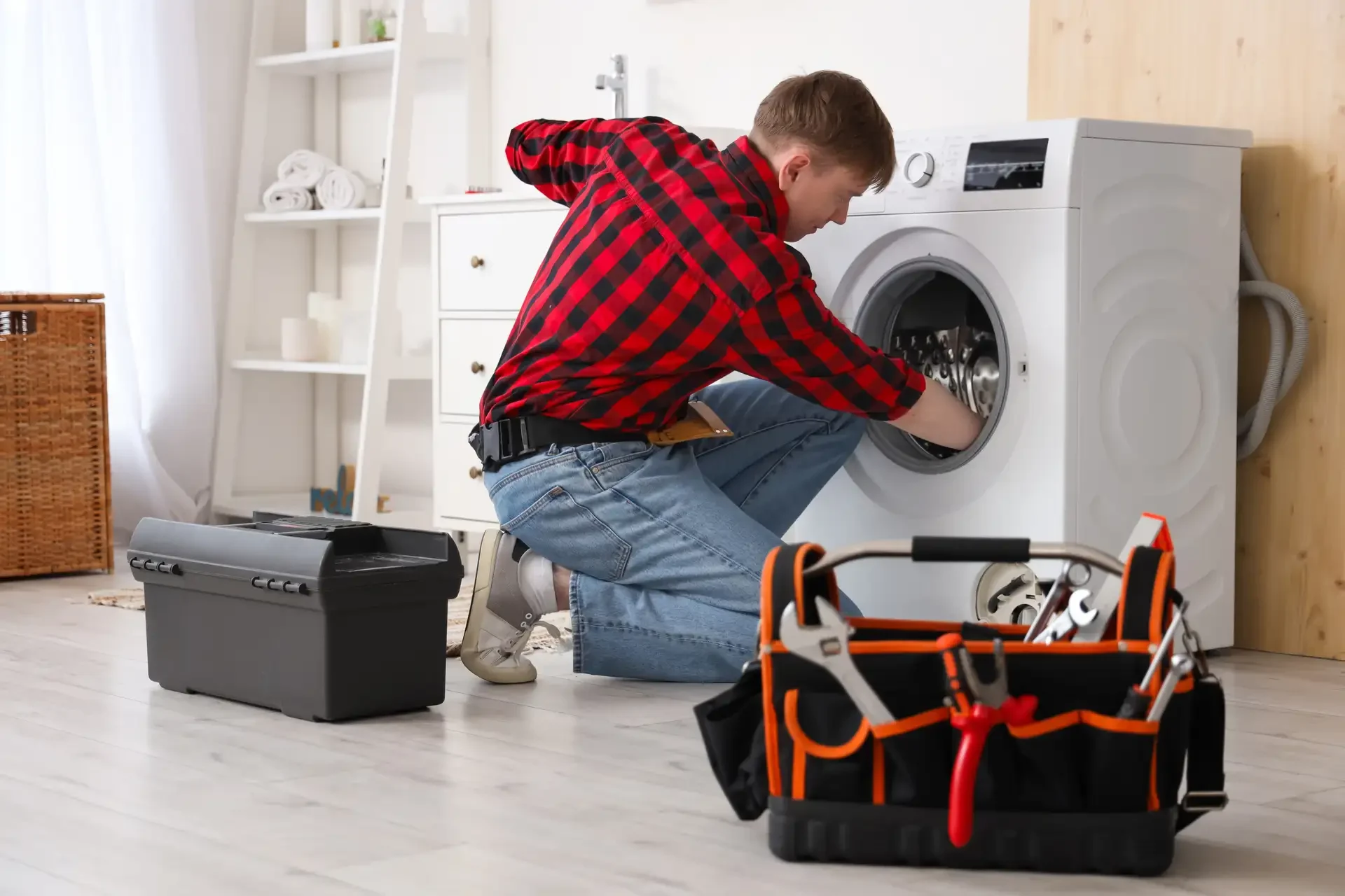 Appliance repair technician in red plaid shirt kneeling beside white front-loading washing machine with hands inside drum, tool bag and black toolbox visible on floor