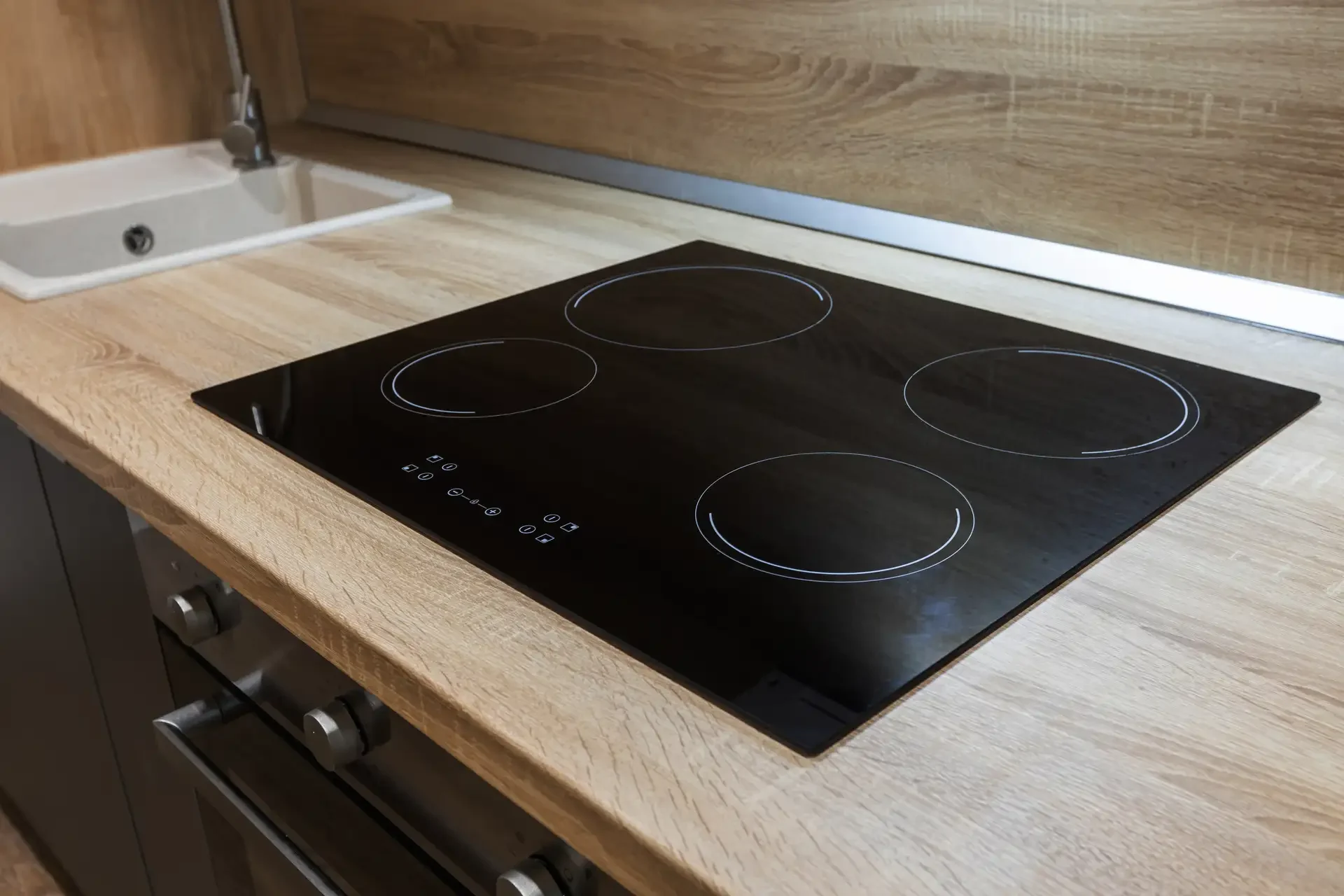 Modern black glass-ceramic electric cooktop with four white-outlined cooking zones integrated into kitchen countertop with built-in oven below