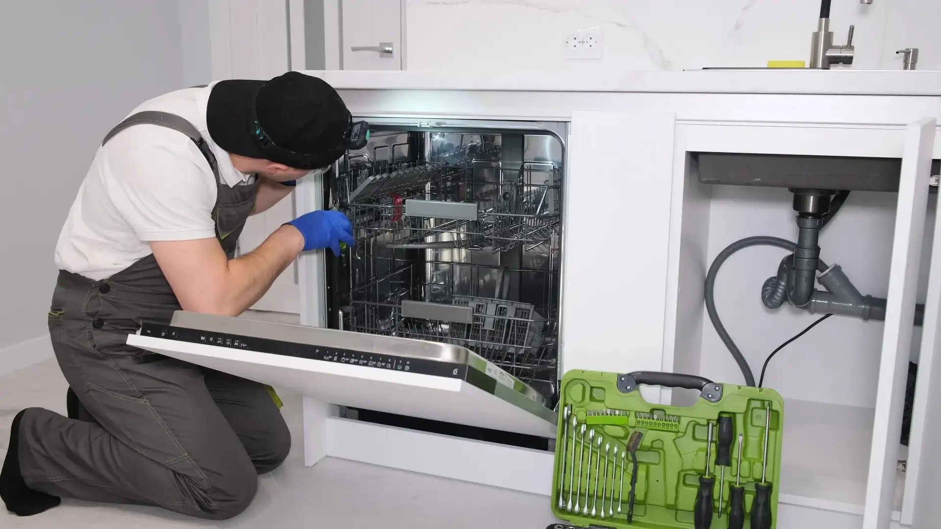 Professional technician in blue gloves and headlamp kneeling to repair open built-in dishwasher with green tool kit visible