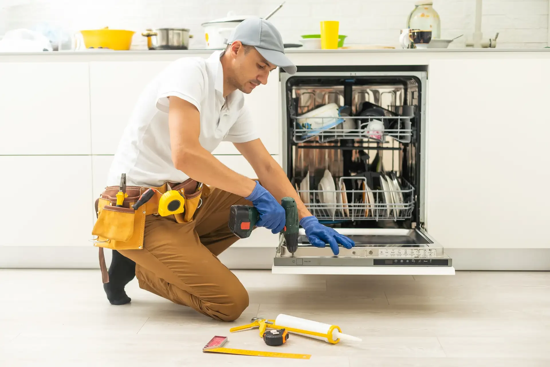 Professional technician kneeling while using power drill to repair built-in dishwasher control panel in residential kitchen