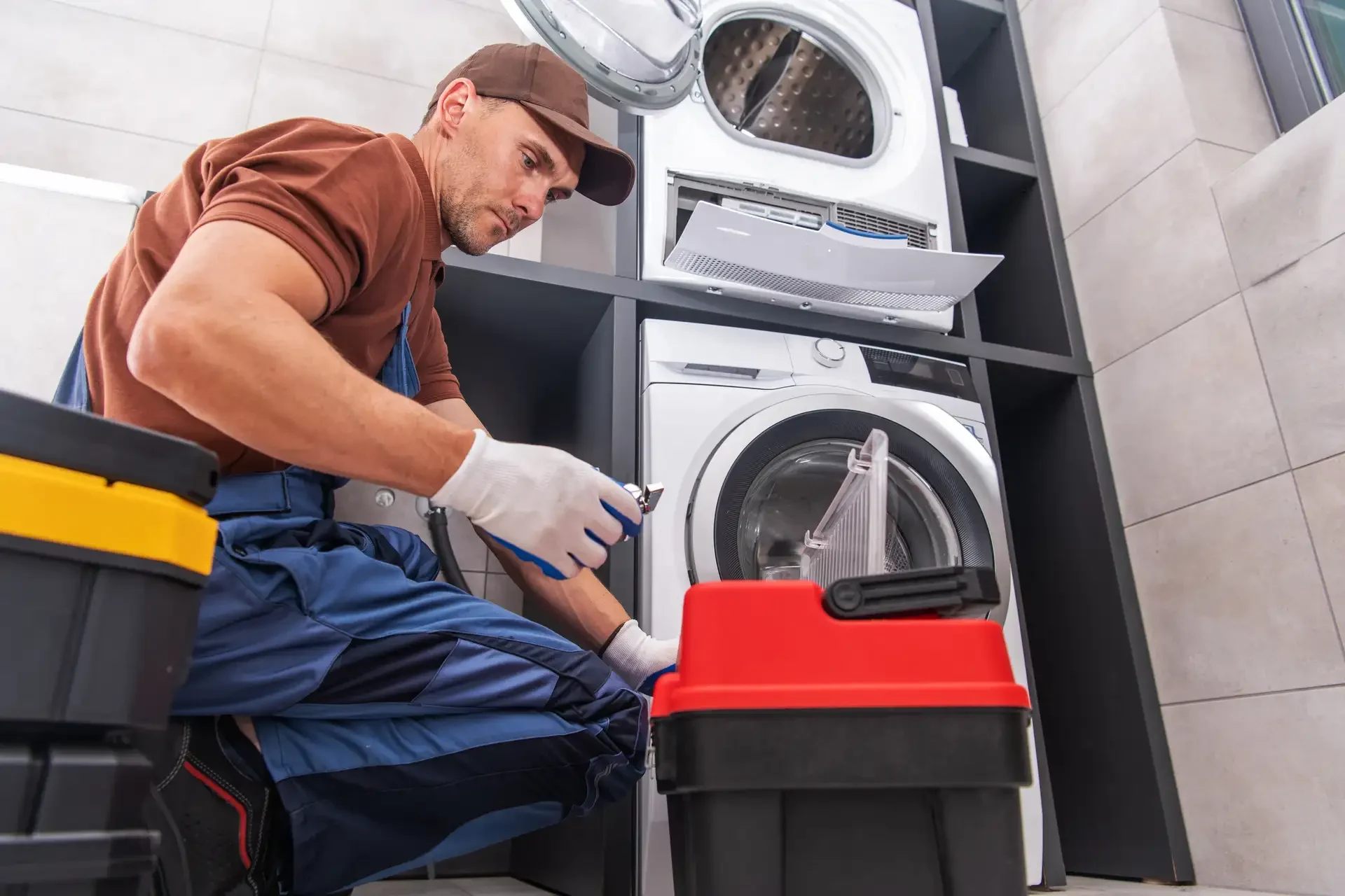 Appliance repair technician in work gloves and overalls using hand tool to repair front-loading washing machine in stacked laundry configuration with toolbox visible