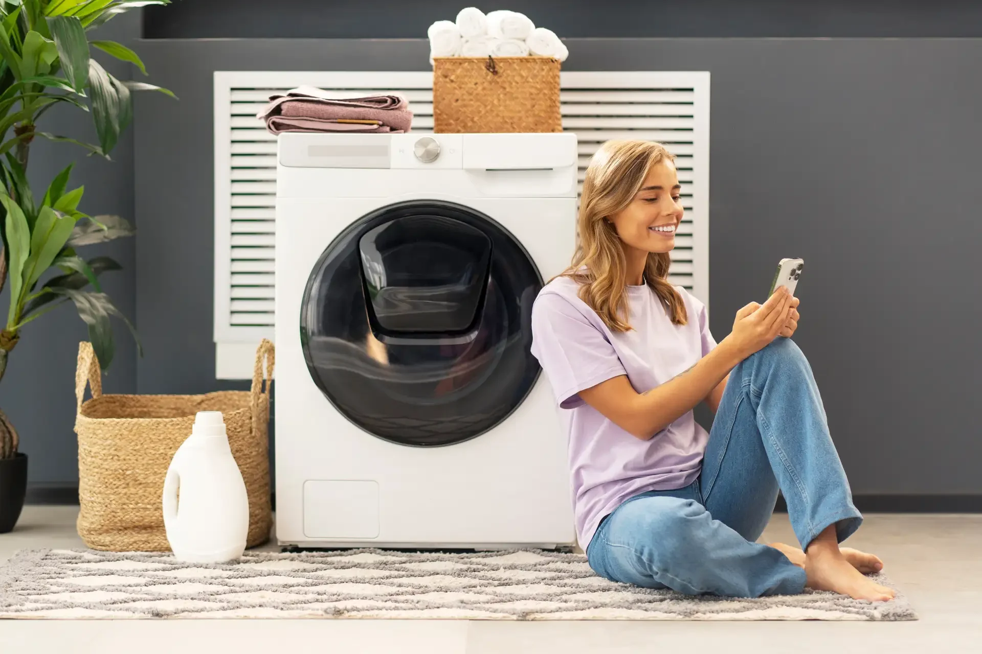 Young woman sitting beside modern front-loading washing machine in organized laundry room, smiling while using smartphone