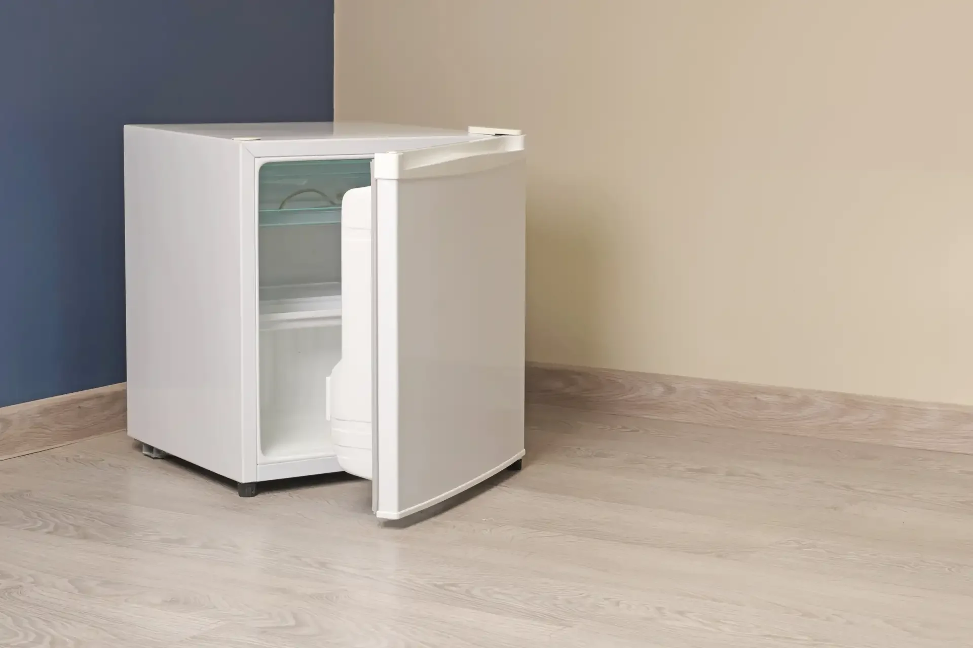 Open mini refrigerator with empty interior showing glass shelf and freezer compartment, positioned against blue and beige wall