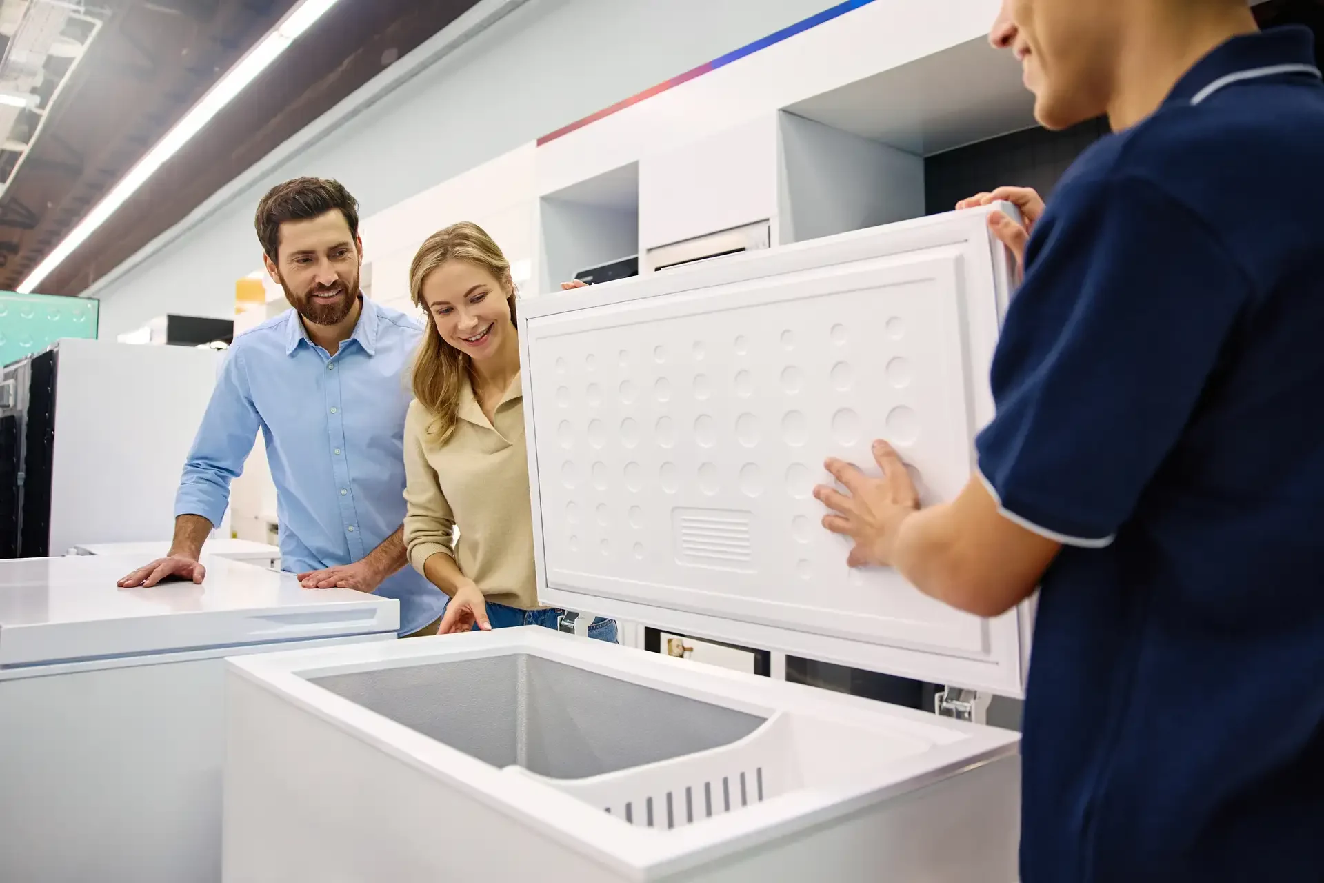 Couple examining open chest refrigerator with store employee in appliance showroom during sales consultation