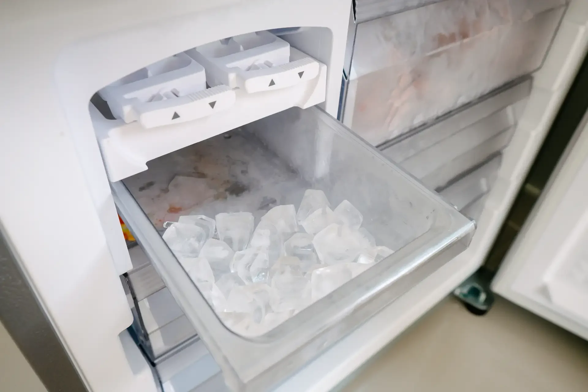 Interior view of refrigerator freezer compartment showing built-in ice maker with ice tray and bin full of clear ice cubes