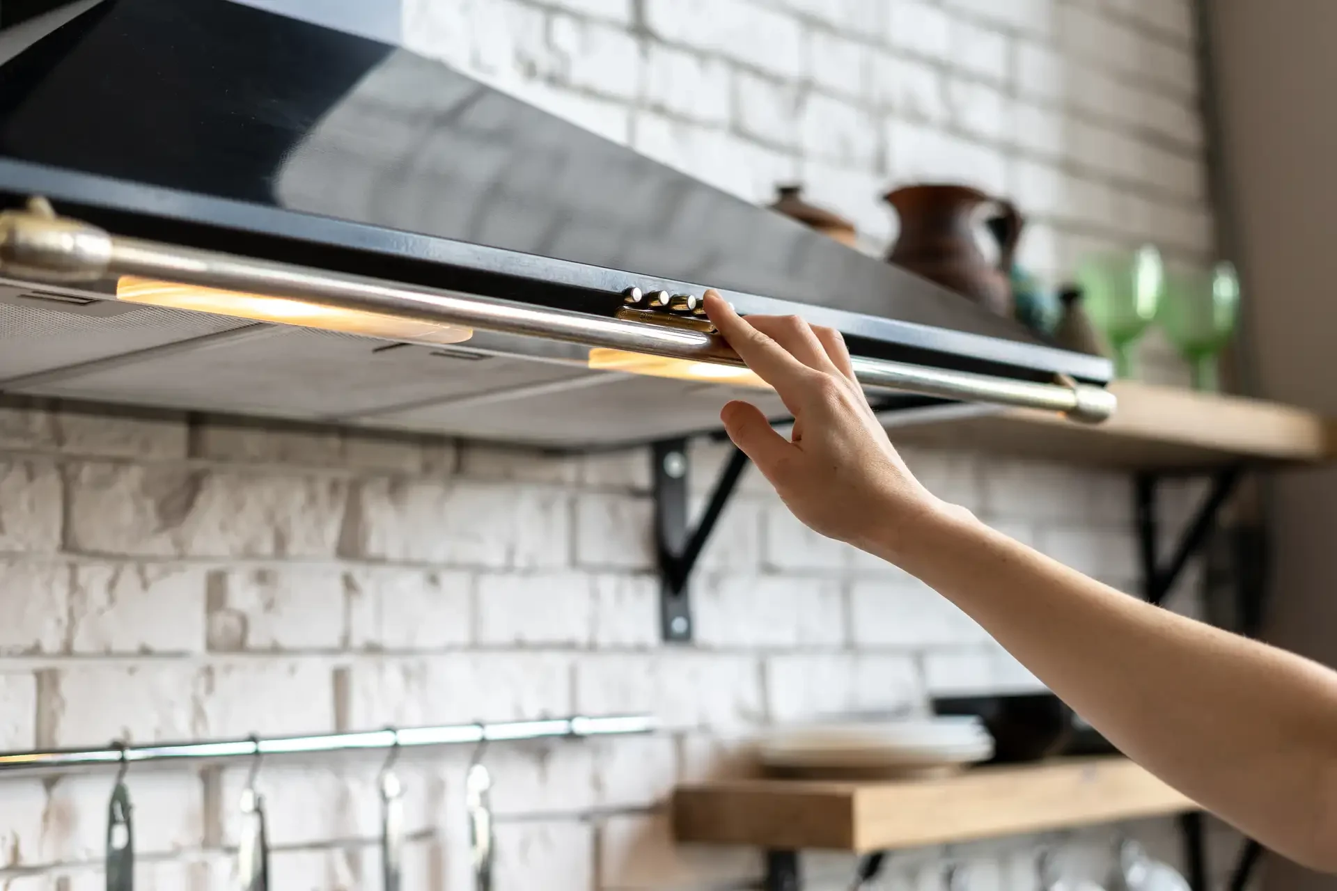 Person operating control panel on illuminated under-cabinet range hood in modern kitchen with white brick backsplash