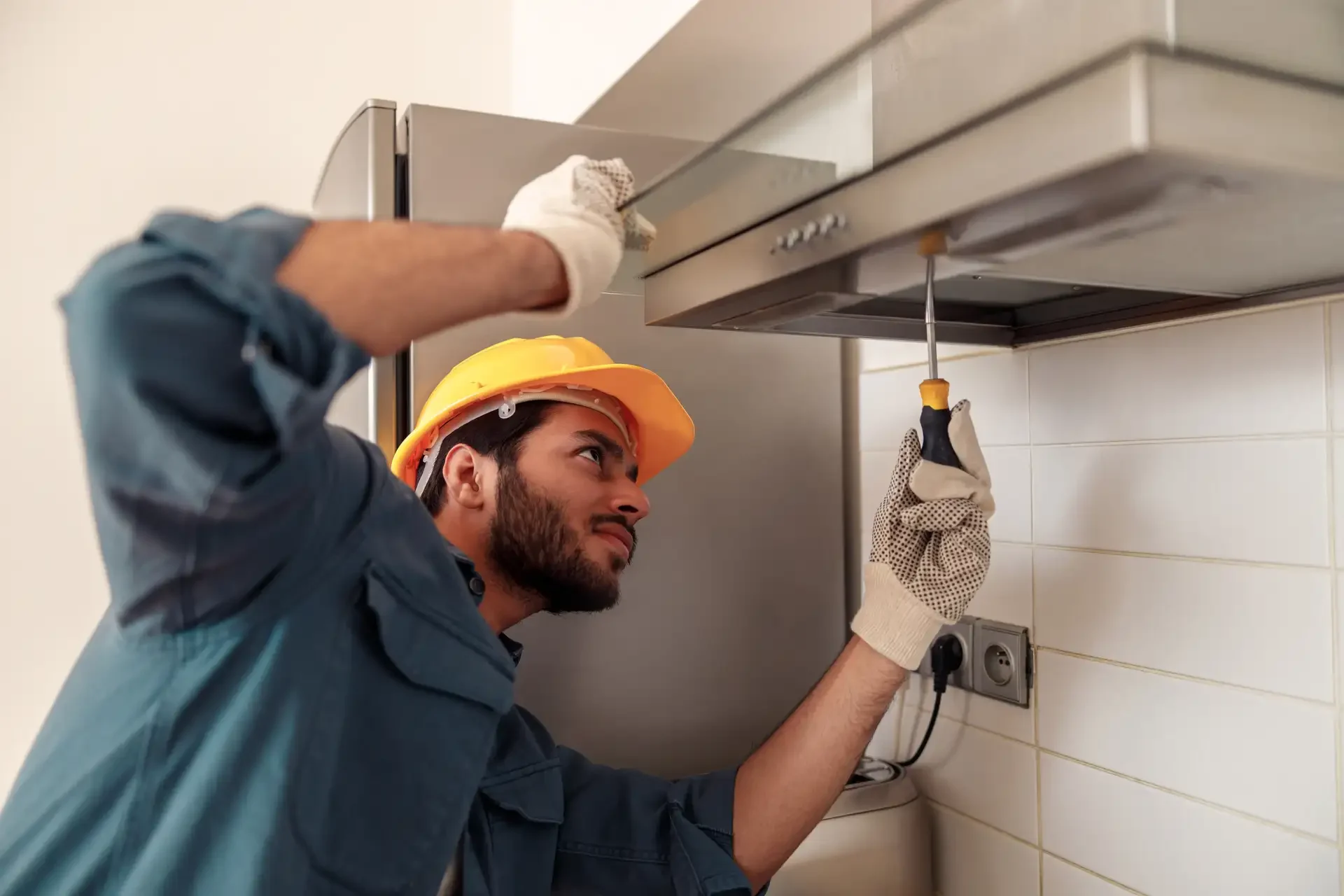 Professional technician wearing hard hat and gloves using screwdriver to repair kitchen range hood with appliance partially opened