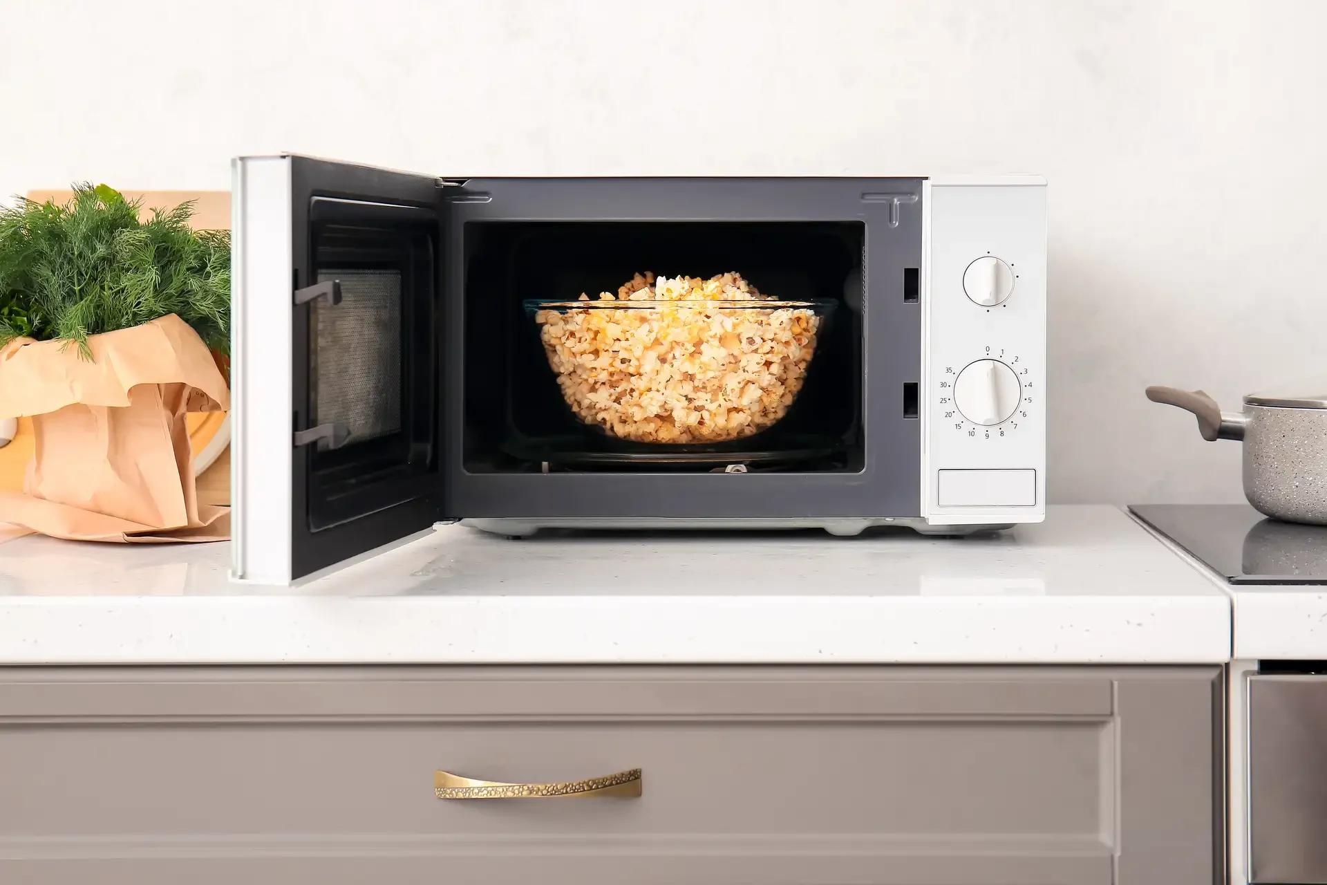 Open countertop microwave with glass bowl of freshly popped popcorn inside, showing white exterior and dial controls in kitchen setting