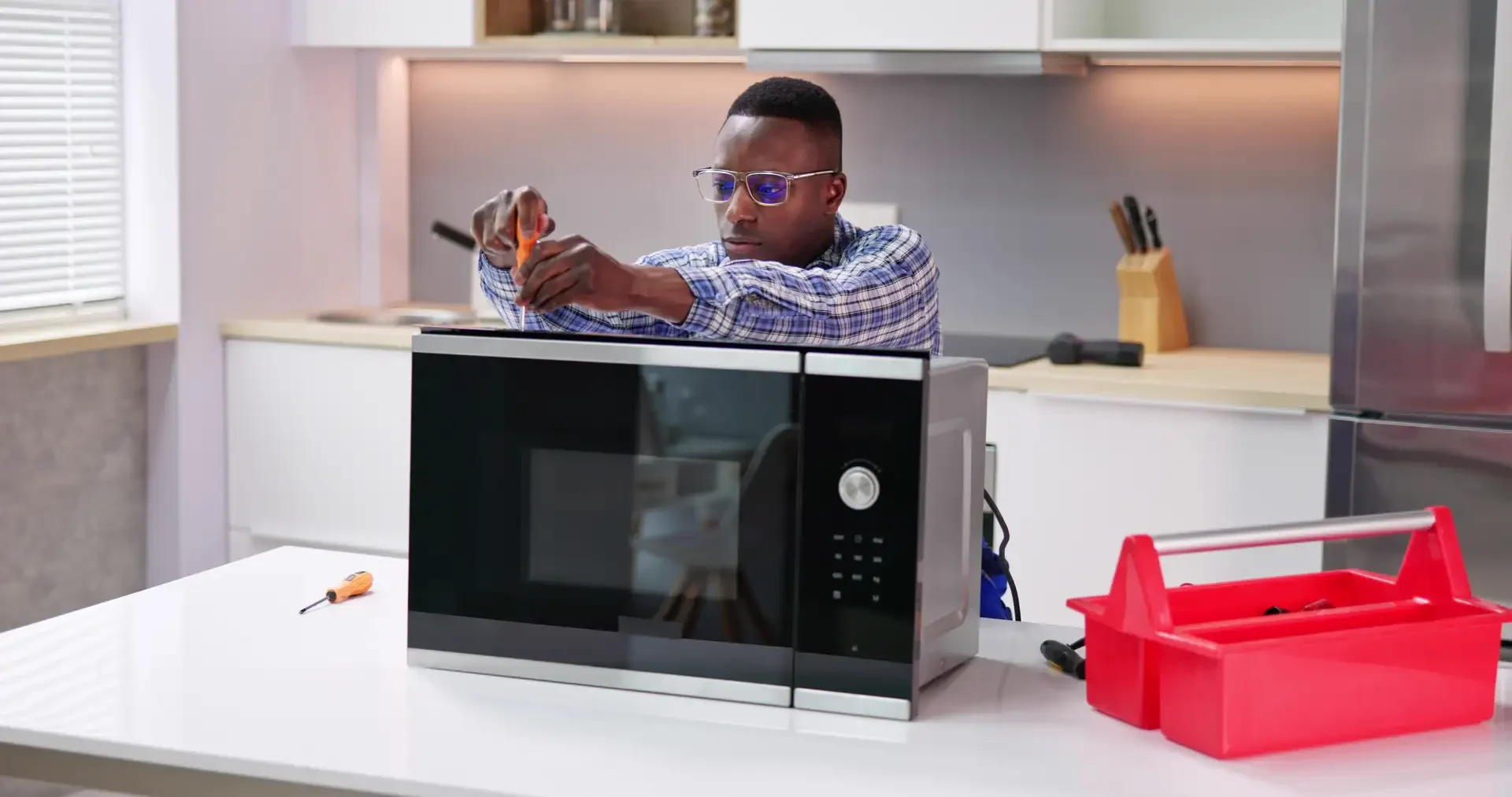 Appliance repair technician using screwdriver to service microwave with open top panel in kitchen, red toolbox and tools nearby