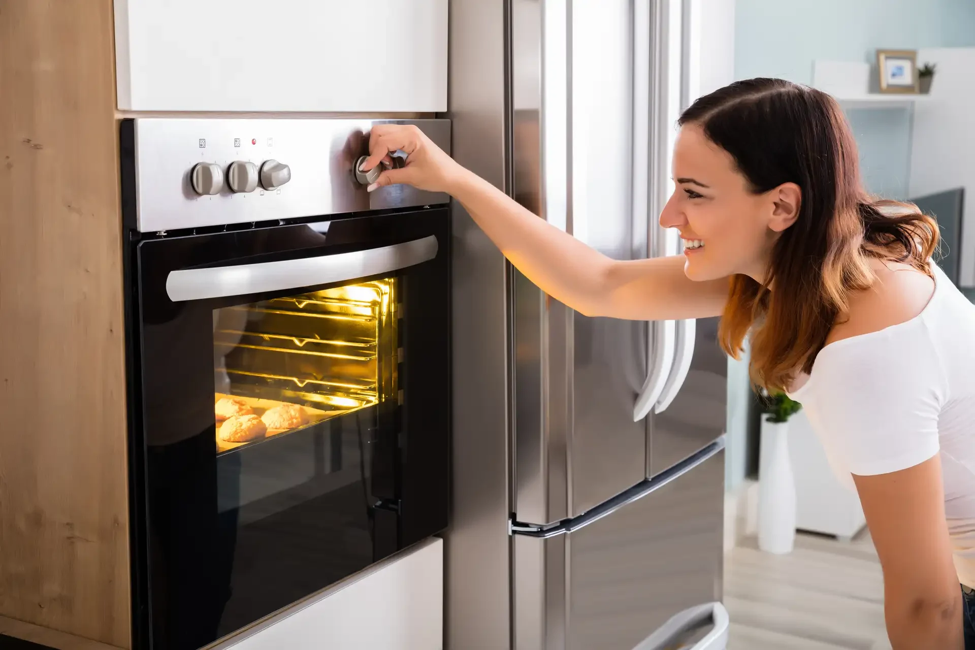 Woman operating a built-in oven in modern kitchen, turning control knob while oven interior is lit and baking food