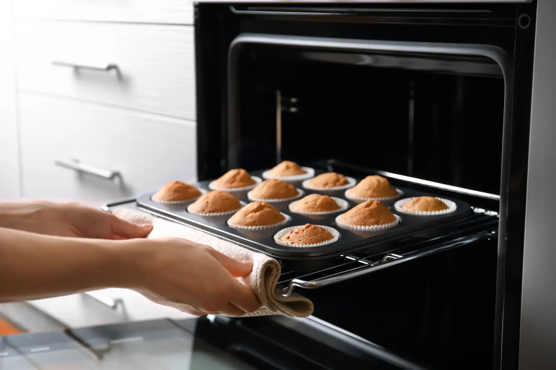 Person removing freshly baked muffins from modern built-in oven using oven mitt in contemporary kitchen