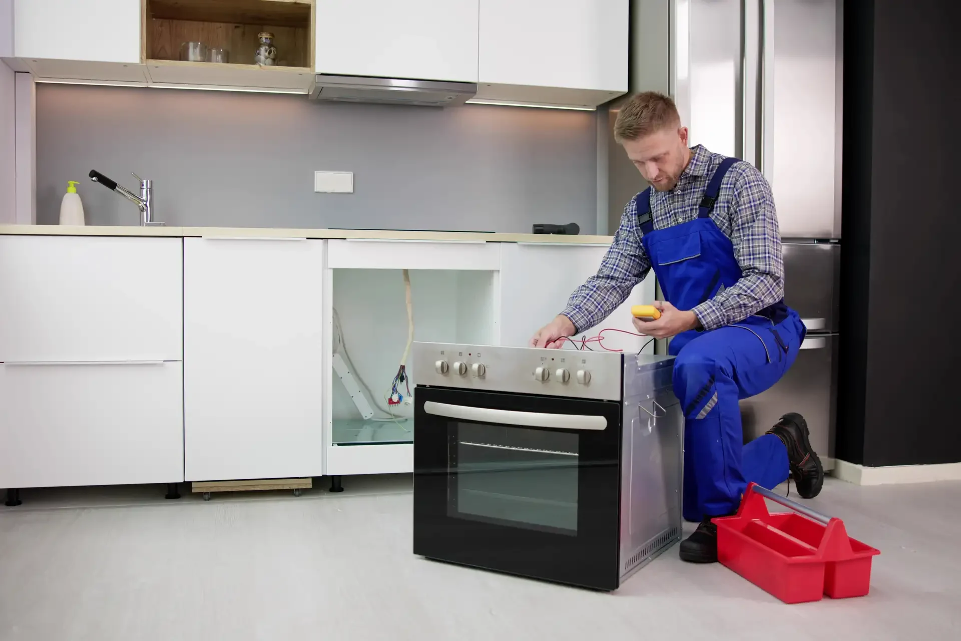 Appliance repair technician in blue uniform using multimeter to diagnose electrical issues on removed oven with exposed cabinet wiring