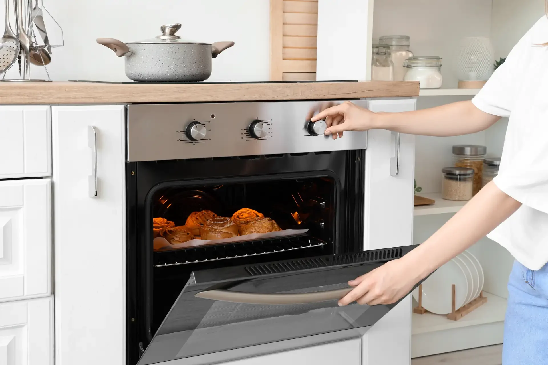 Modern built-in oven with open door showing cinnamon rolls baking inside, person adjusting control knob