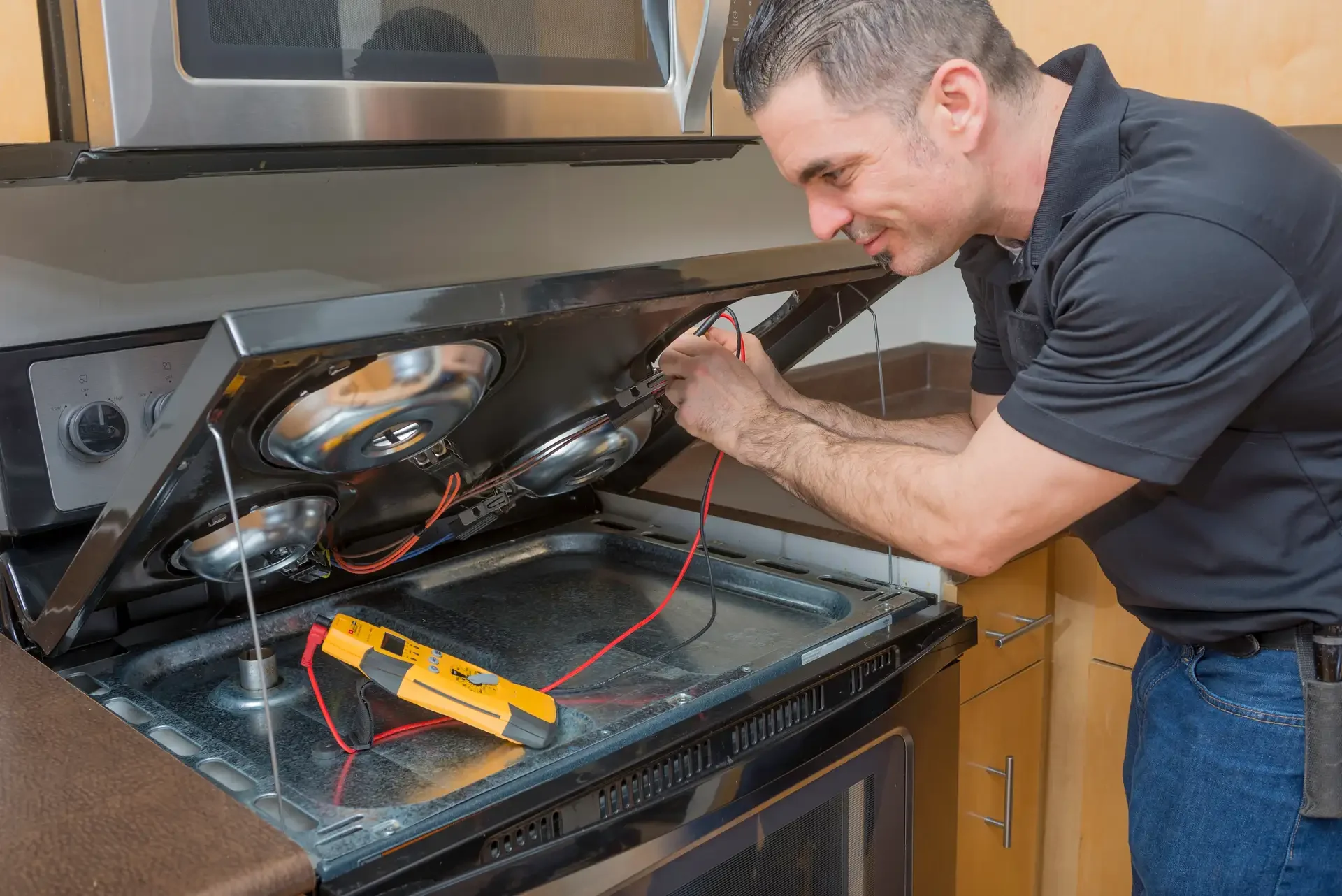 Technician repairing electric oven with cooktop lifted open, exposing internal wiring and components, multimeter on surface for electrical diagnostics