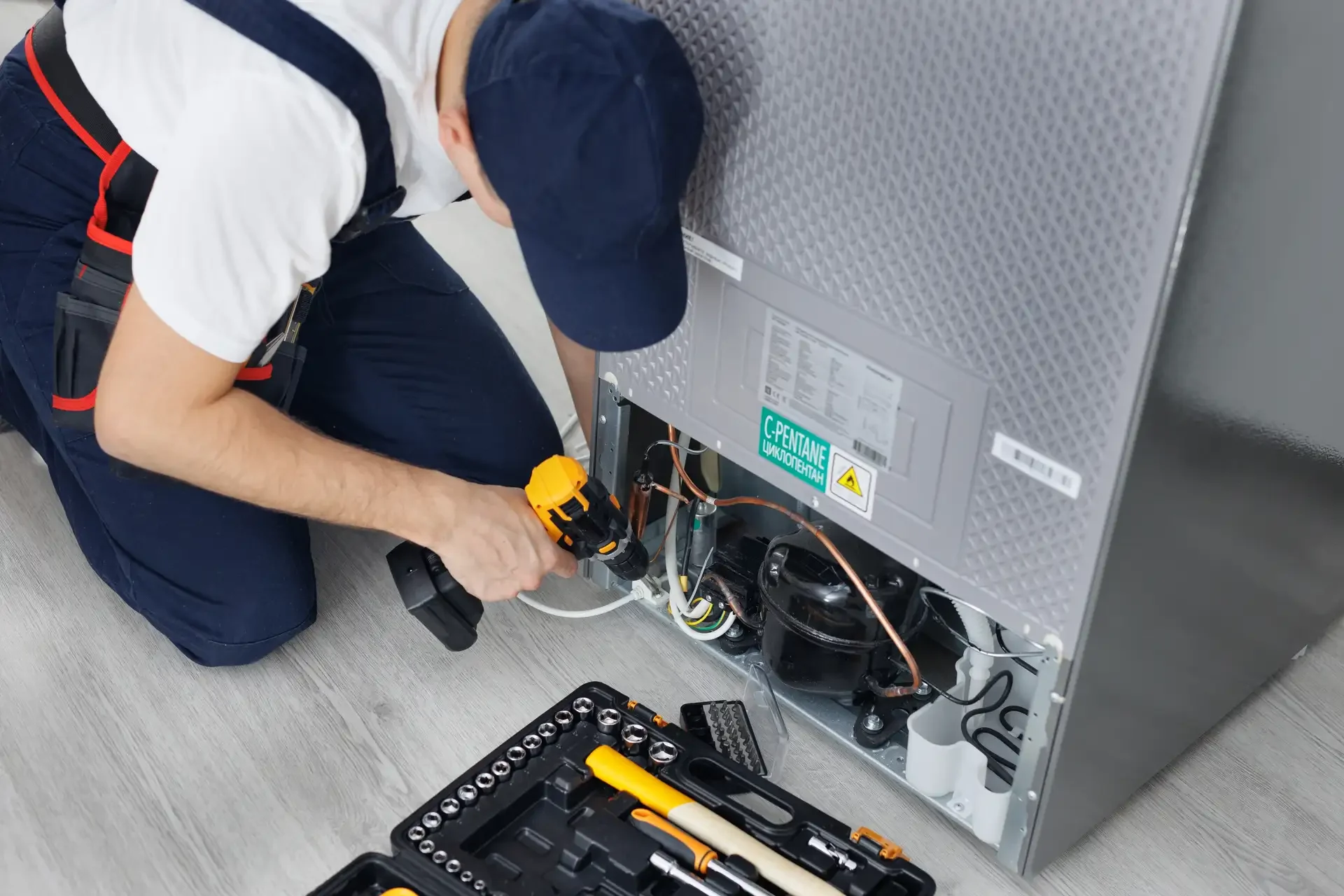 Technician kneeling beside refrigerator using cordless drill on exposed compressor area with open back panel, repair toolkit on floor