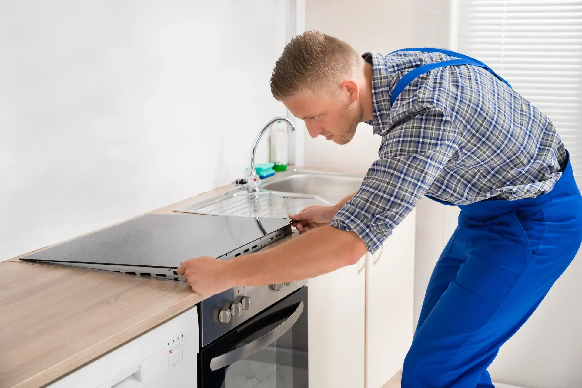 Appliance repair technician in blue overalls working on electric oven, removing glass cooktop in kitchen setting