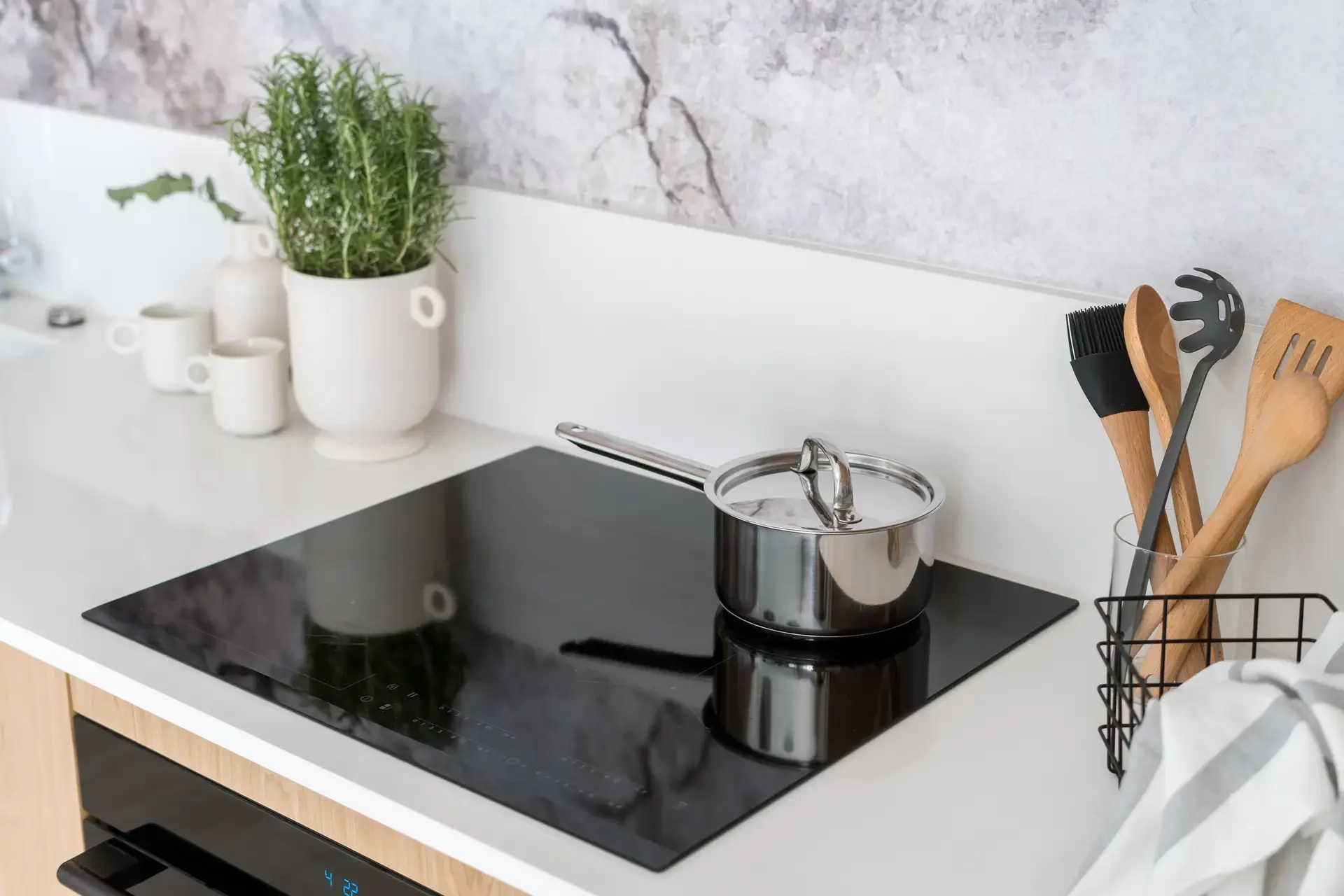 Modern black glass induction cooktop with stainless steel saucepan on clean kitchen counter surrounded by cooking utensils and herbs