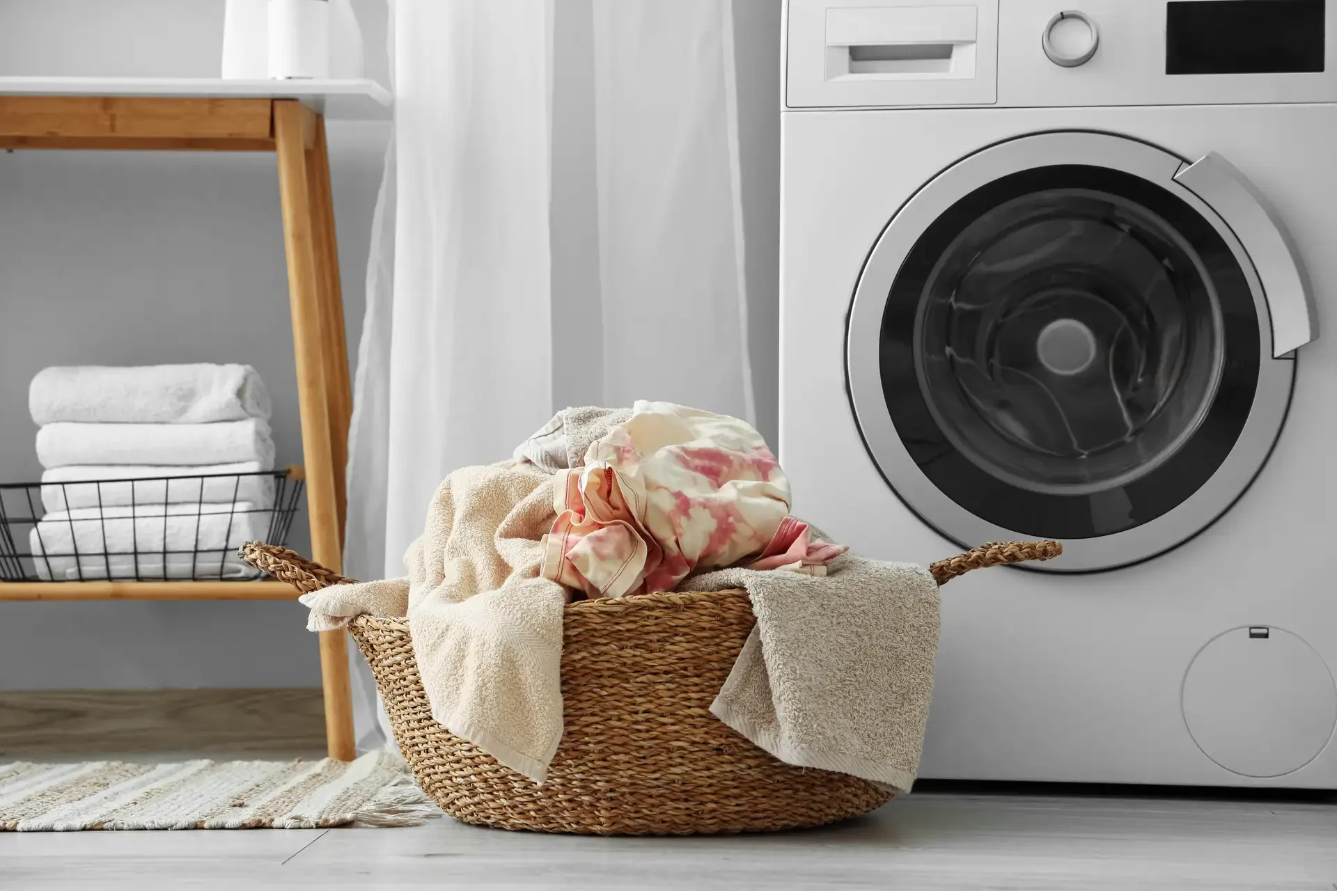 Front-loading washing machine in organized laundry room with wicker basket of clean laundry and folded towels on wooden stand