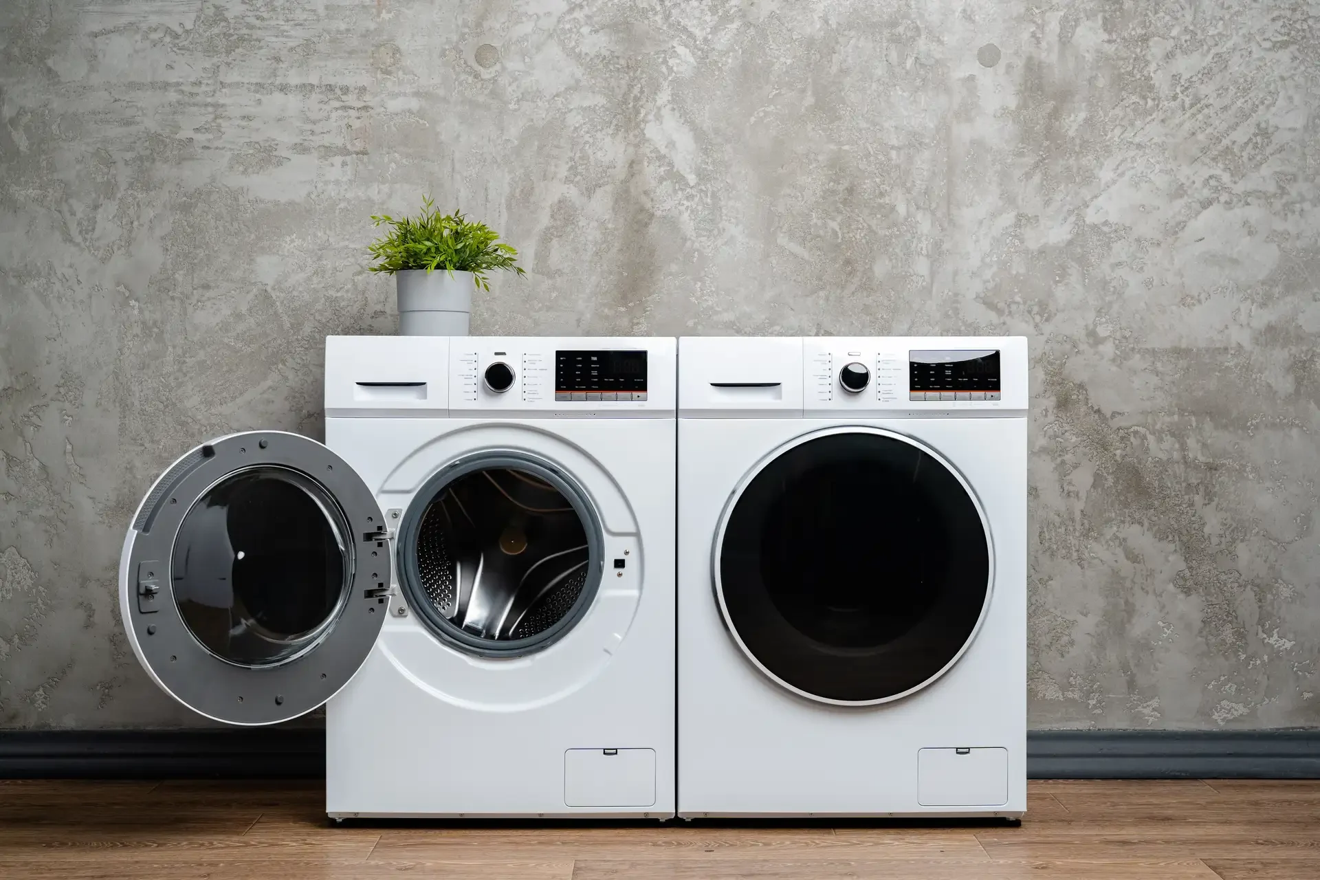 Modern front-loading washing machine with open door showing stainless steel drum, positioned next to matching dryer against concrete wall