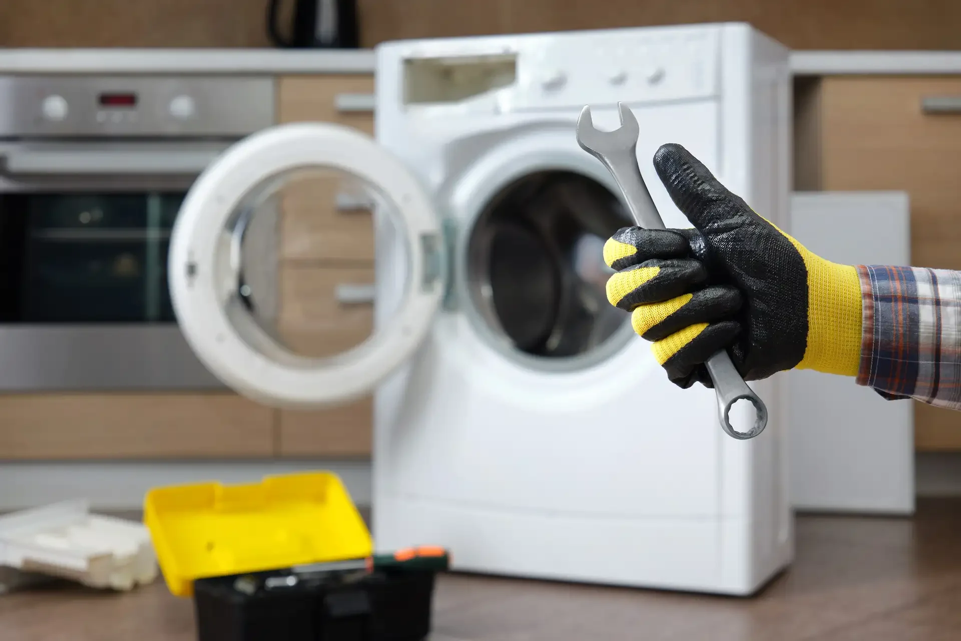 Technician's gloved hand holding wrench during washing machine repair, with open front-loading washer and toolbox on floor