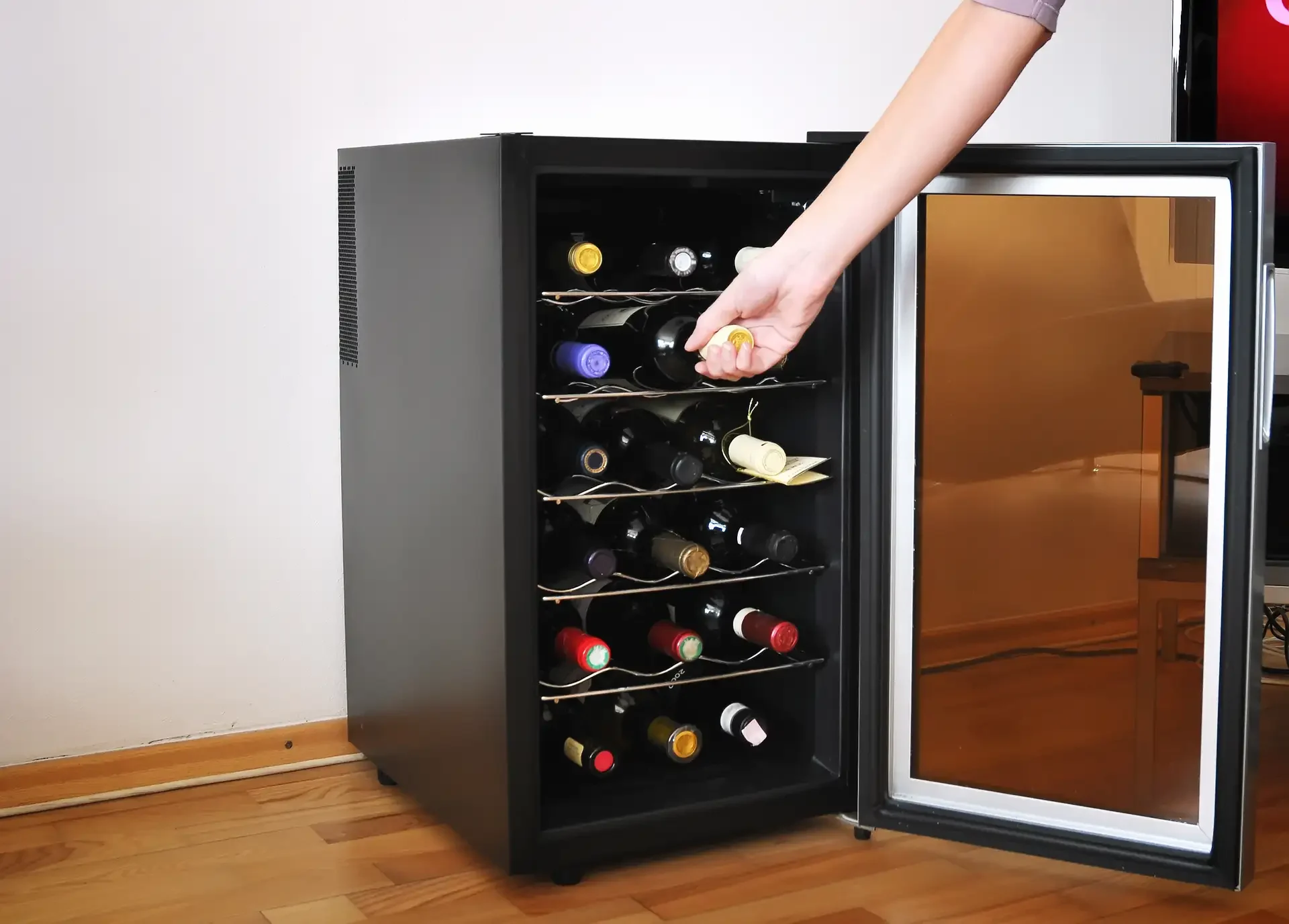 Person reaching into an open wine refrigerator with multiple shelves stocked with wine bottles in a residential setting